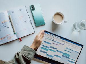 scrivania vista dall'alto con mano di donna che regge un tablet con aperta una schermata calendario