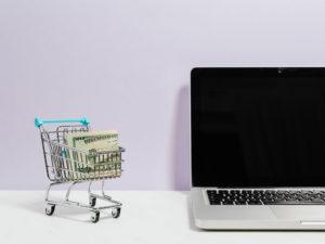 laptop e piccolo carrello della spesa con banconote sopra una scrivania bianca